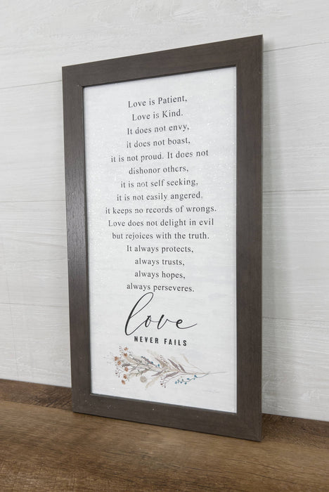 A framed typographic wall art print with a dark wood frame, featuring a quote about love and the word 'love' in script with small floral accents near the bottom, shown leaning against a white paneled wall on a wooden surface.
