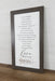 A framed typographic wall art print with a dark wood frame, featuring a quote about love and the word 'love' in script with small floral accents near the bottom, shown leaning against a white paneled wall on a wooden surface.